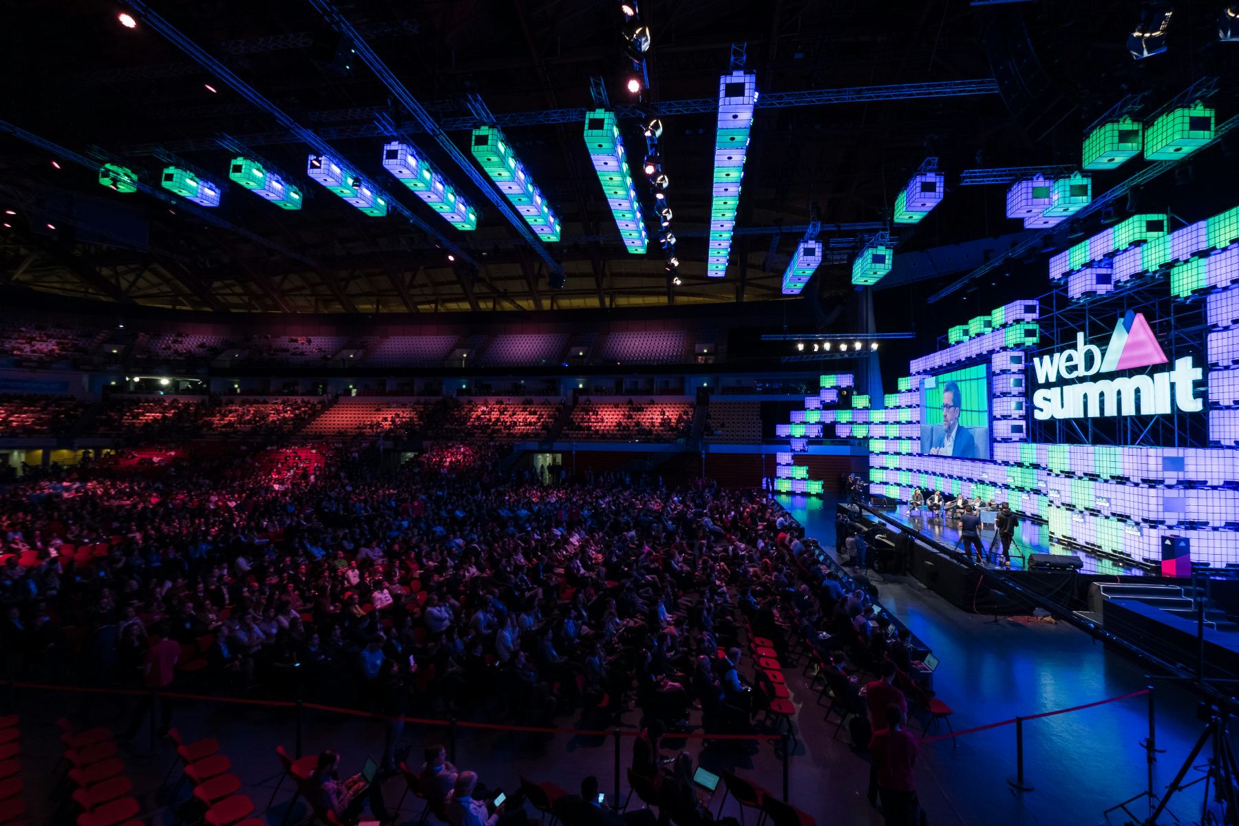 An arena-scale summit stage with a lit backdrop and thousands of seated attendees.