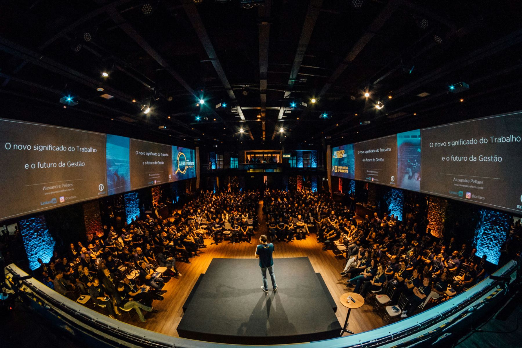 A speaker alone on a central stage surrounded by a full auditorium, with screens on every wall.