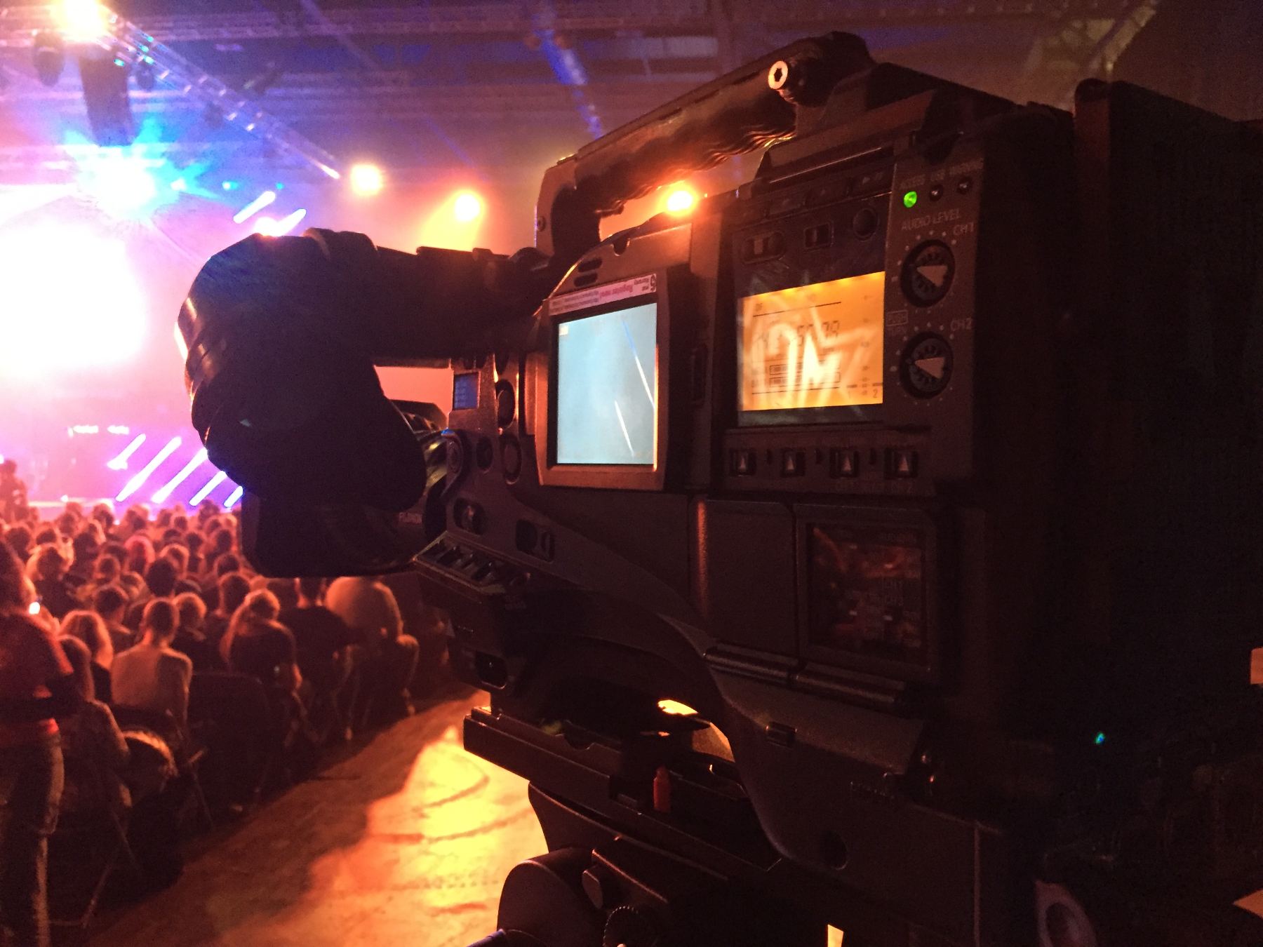 A camera operator shooting a packed standing audience under stage lighting.