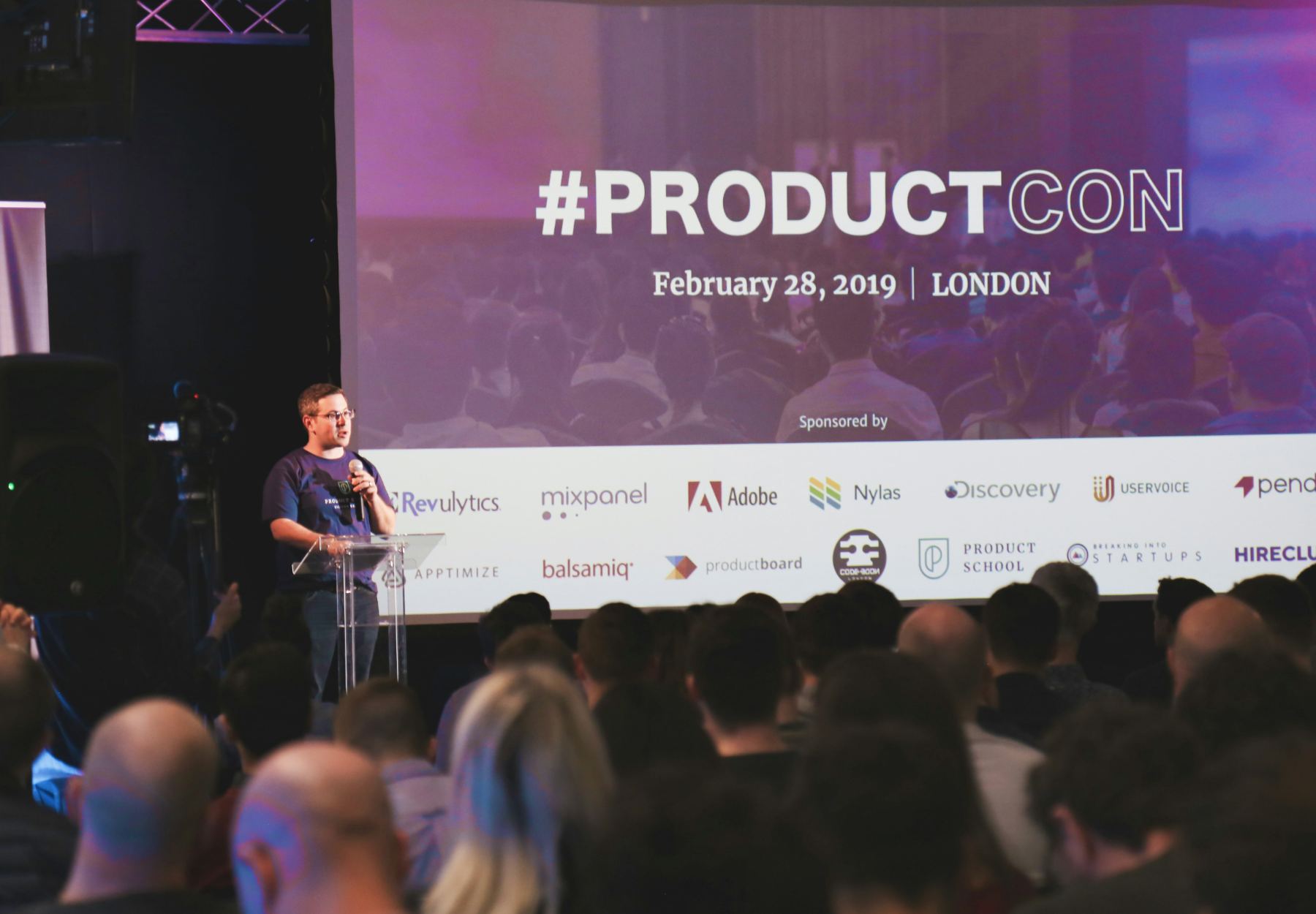 A host speaking from a lectern in front of a seated conference audience and a sponsor screen.