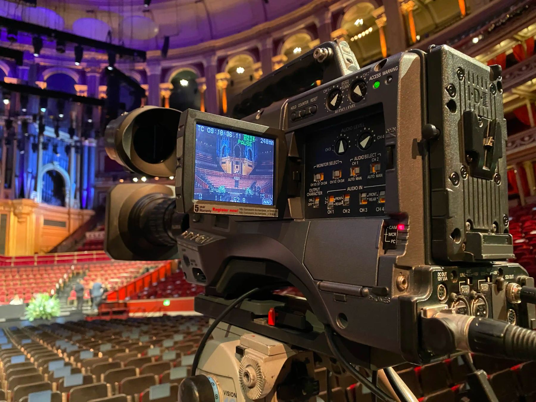 A broadcast camera on the stage of a domed concert hall during rehearsal, timecode running on its monitor.
