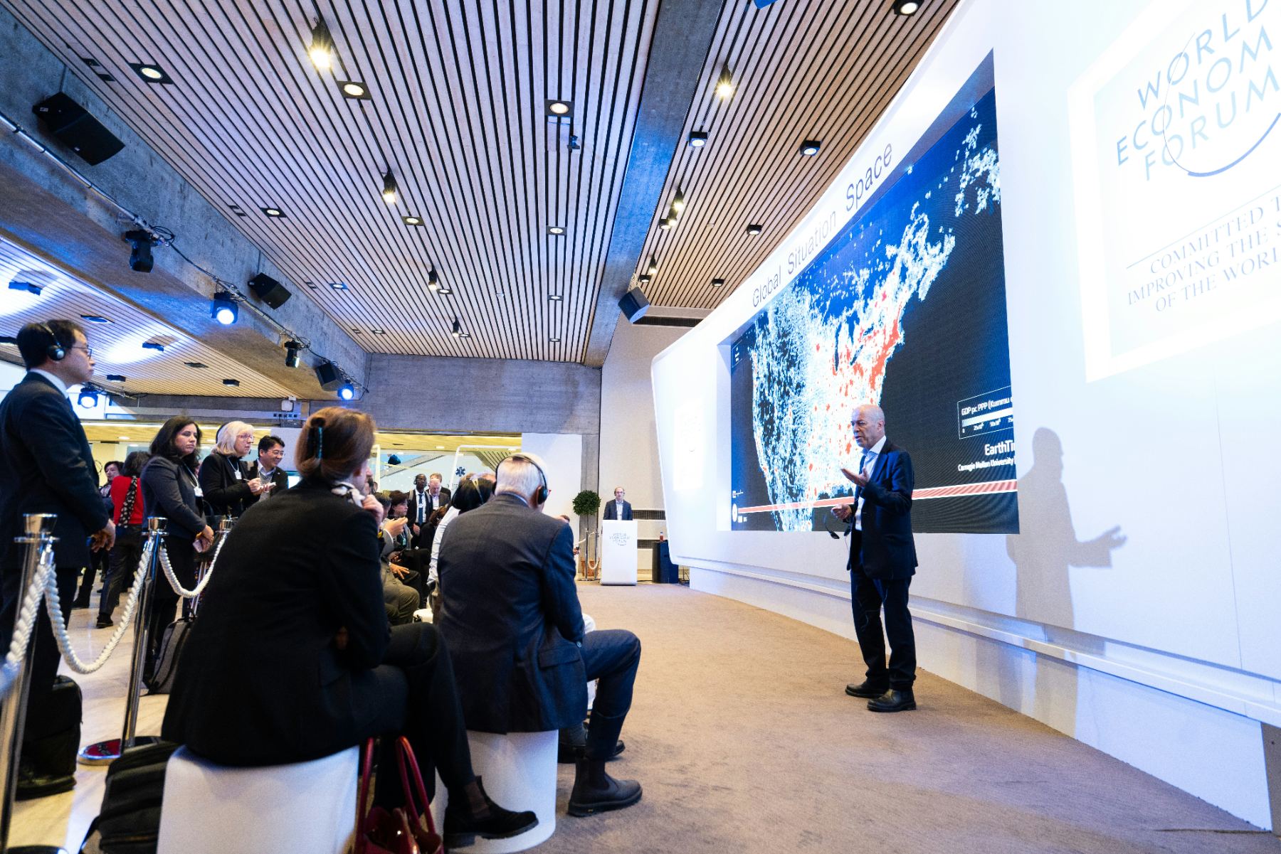 A briefing-room session with a presenter at a data screen and delegates listening on headsets.