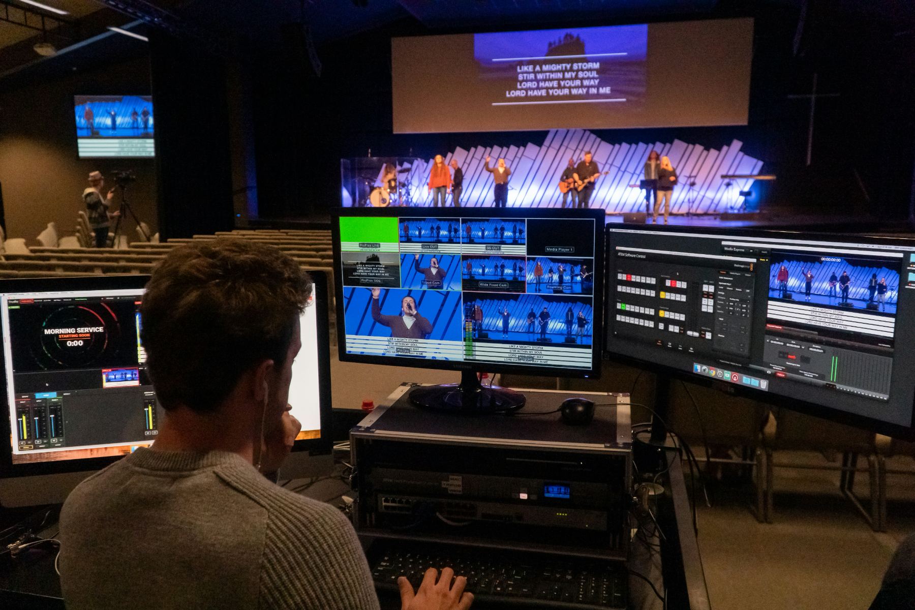 Three crew working side by side in a broadcast gallery, facing a wall of monitors and a lit control desk.