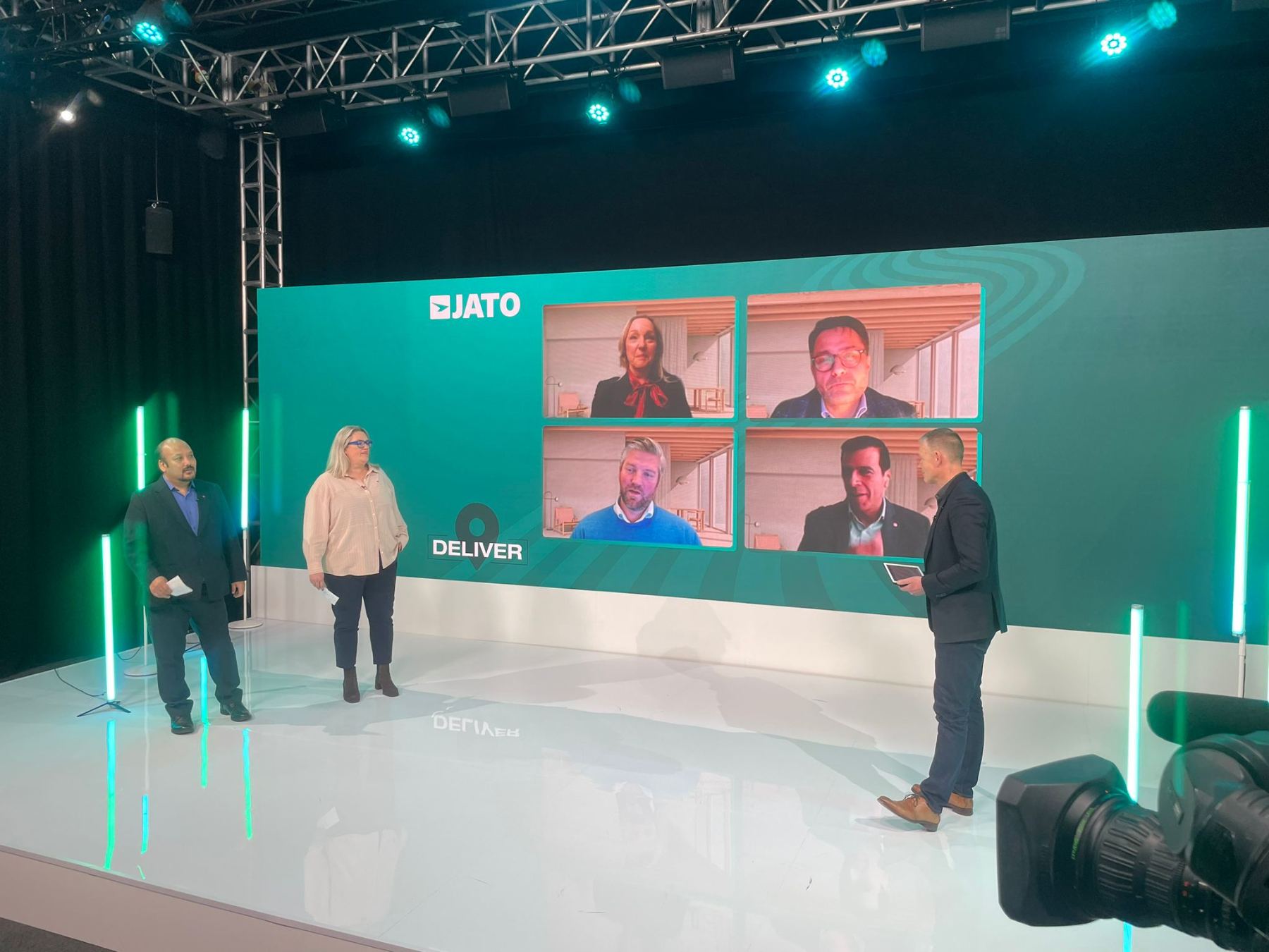 A hybrid studio panel: two presenters on a lit stage beside a large screen of four remote speakers.