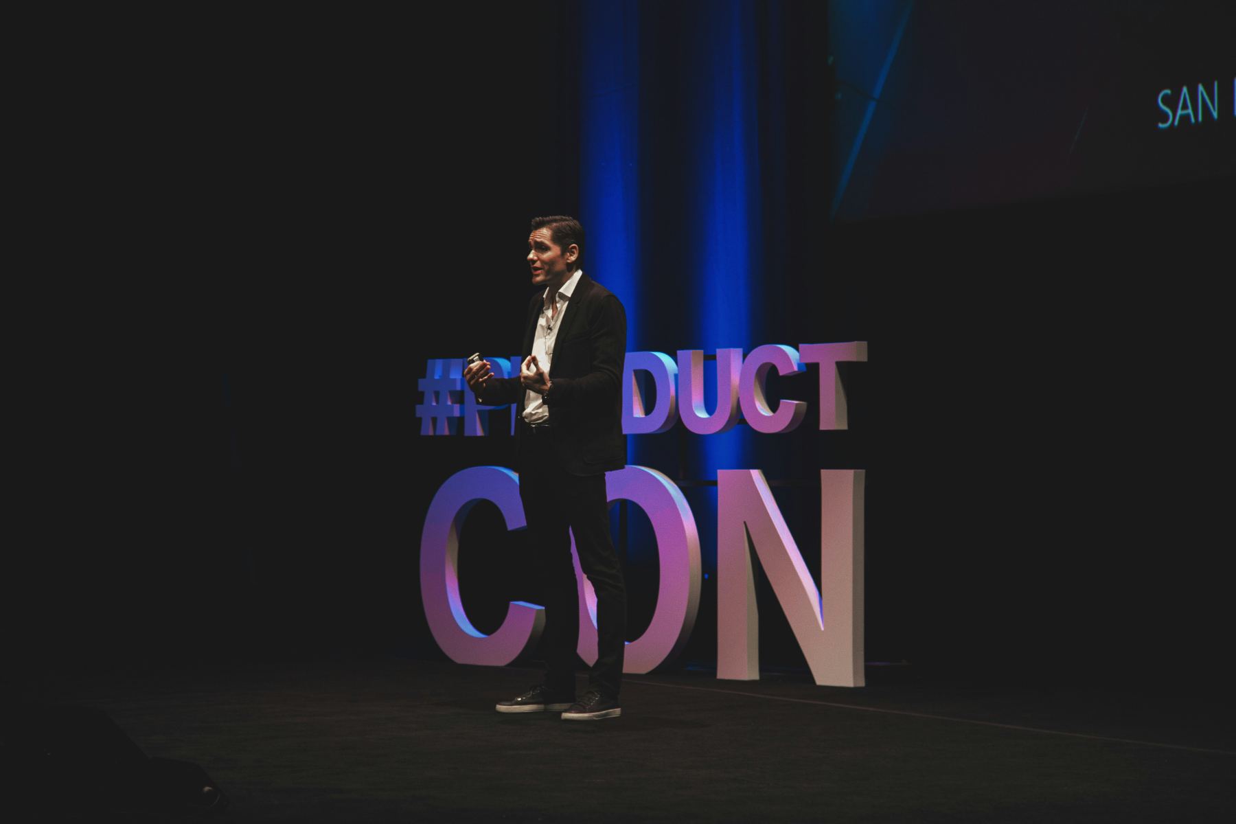 A speaker mid-talk on a dark main stage in front of large three-dimensional conference lettering.